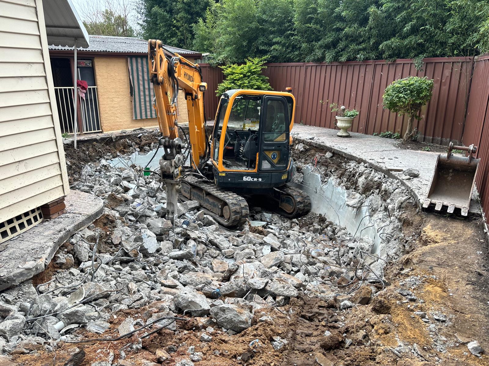 Excavator sitting inside a demolished pool cavity surrounded by broken concrete and steel